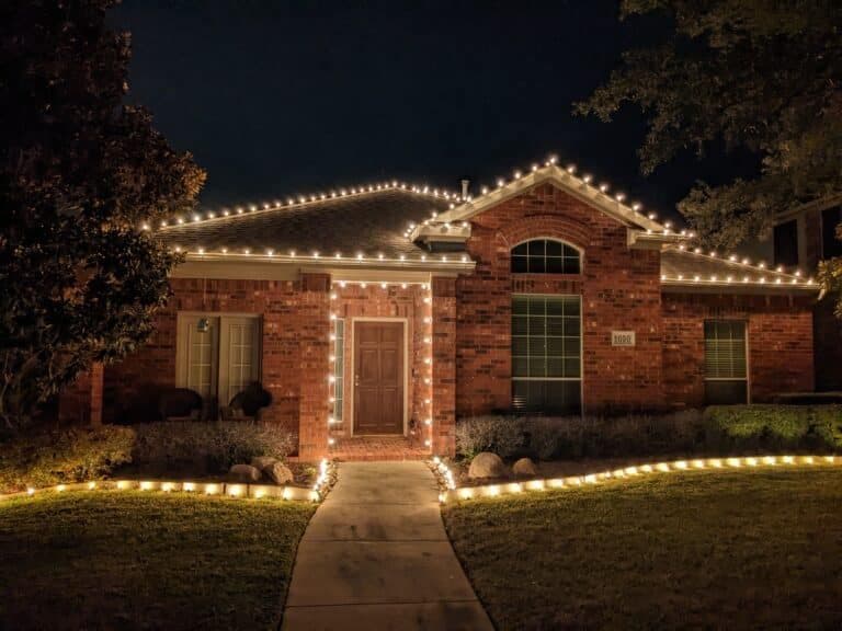 Festively lit brick house at night with white string lights adorning roof and pathway.