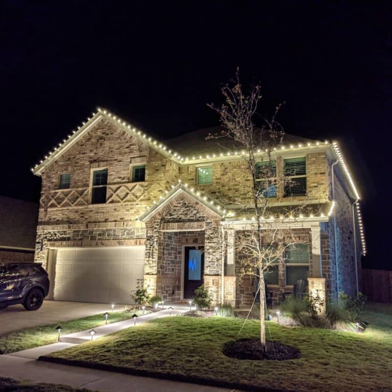 Modern brick house with festive white lights, illuminated at night. Well-maintained lawn.