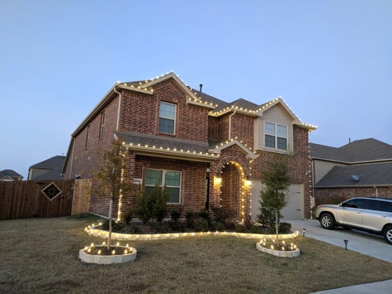 Brick house adorned with Christmas lights and landscaped yard on a clear evening.