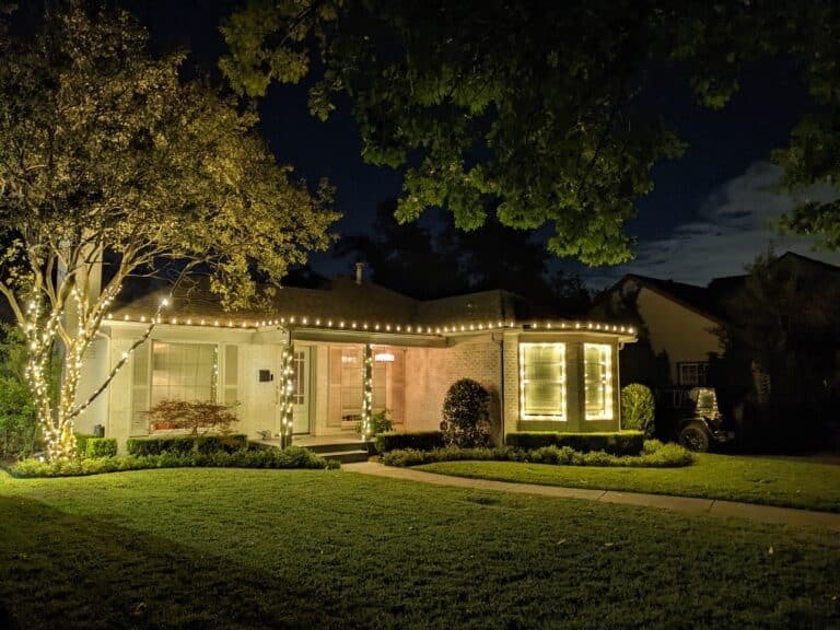 Brightly lit home with string lights and lush green lawn at night. Cozy suburban ambiance.