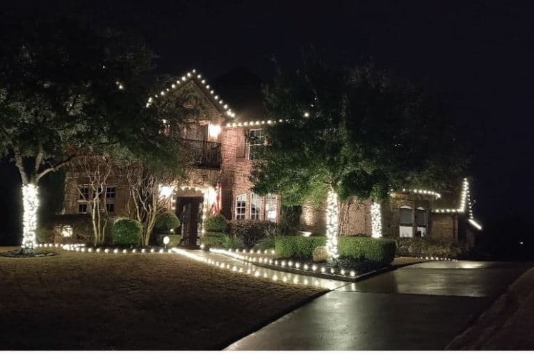 Elegant brick house decorated with festive white lights, surrounded by lush greenery at night.