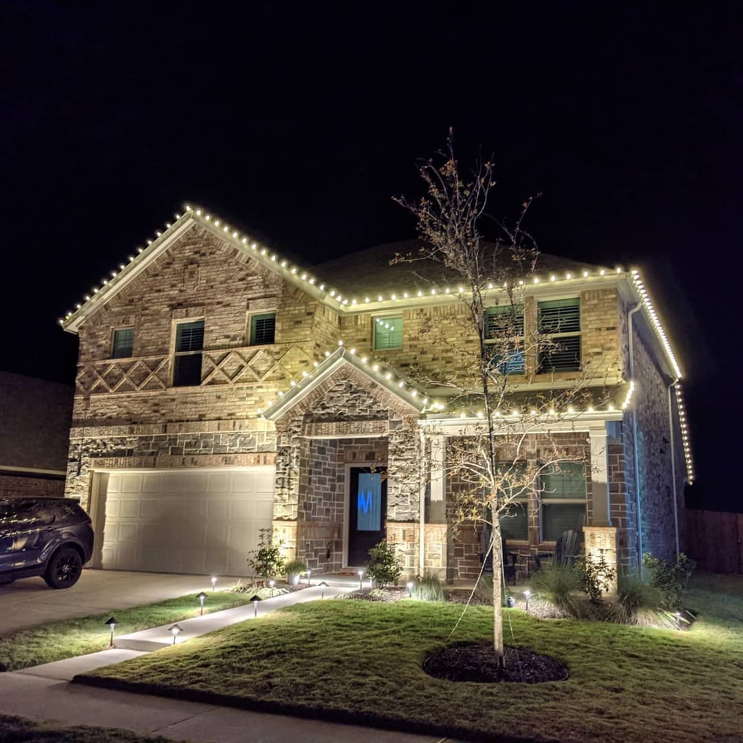 Modern stone house adorned with festive LED lights at night. Lush lawn and pathway lighting visible.