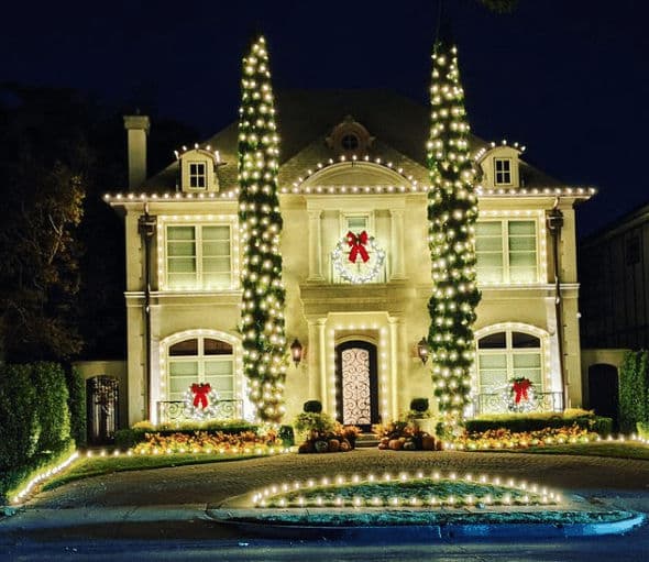 Elegant Christmas-lit house with wreaths and bows, featuring tall festive trees at night.
