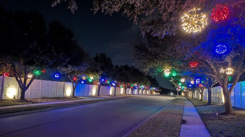 Colorful holiday lights adorn trees along a quiet, empty street at night.