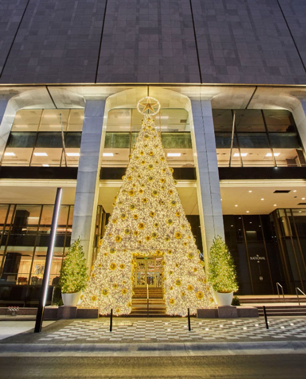 Illuminated Christmas tree adorned with gold ornaments outside a modern building entrance.